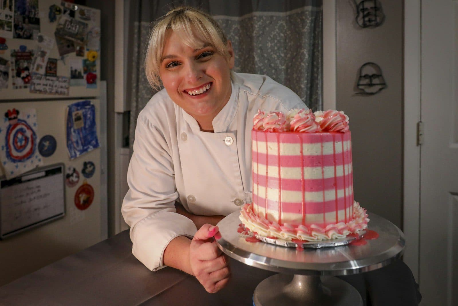 A baker in her whites smiling beside a striped pink layer cake on a turntable.