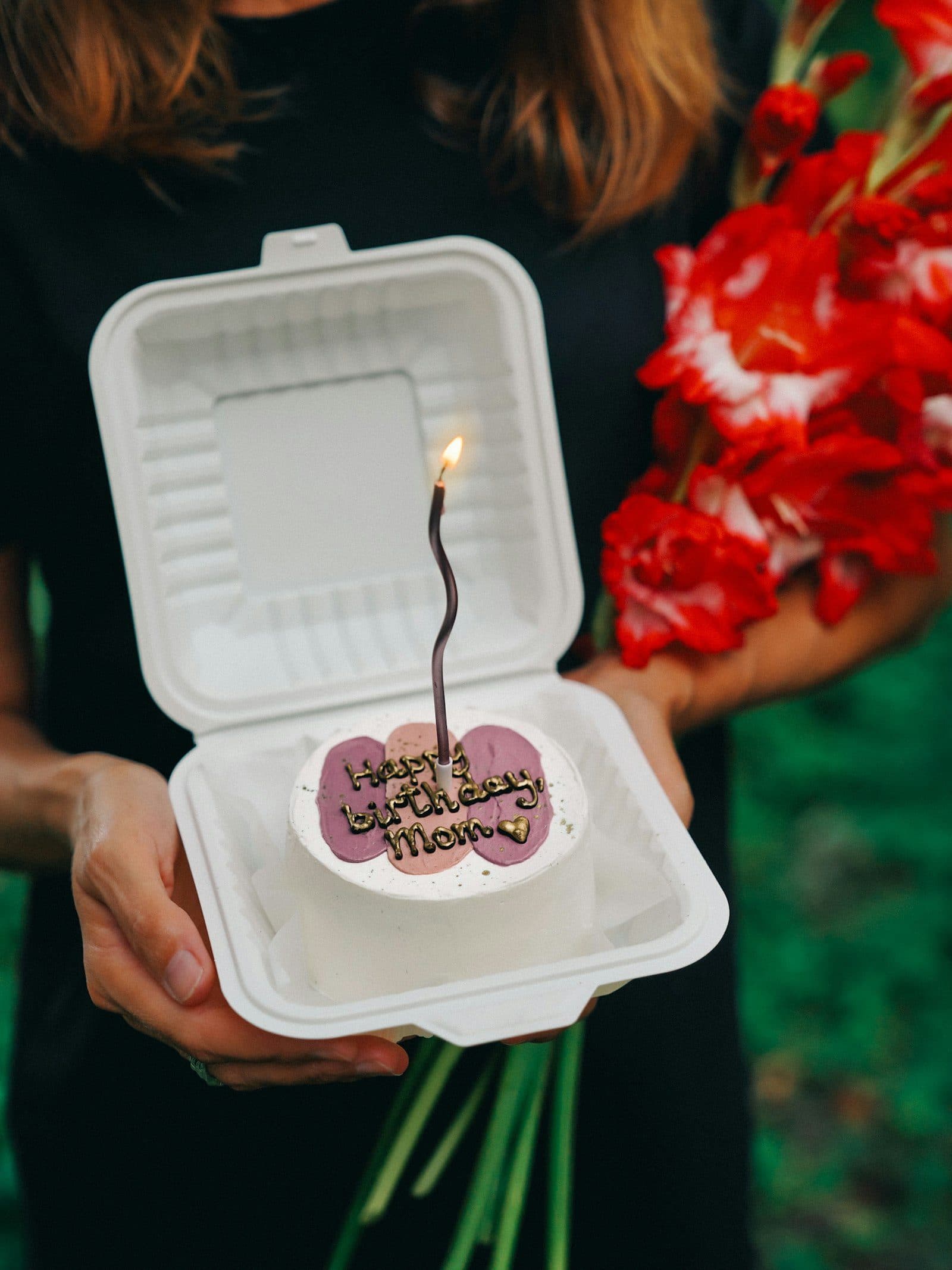 Someone holding out a small birthday cake in an open box, one candle lit, flowers tucked under their arm.