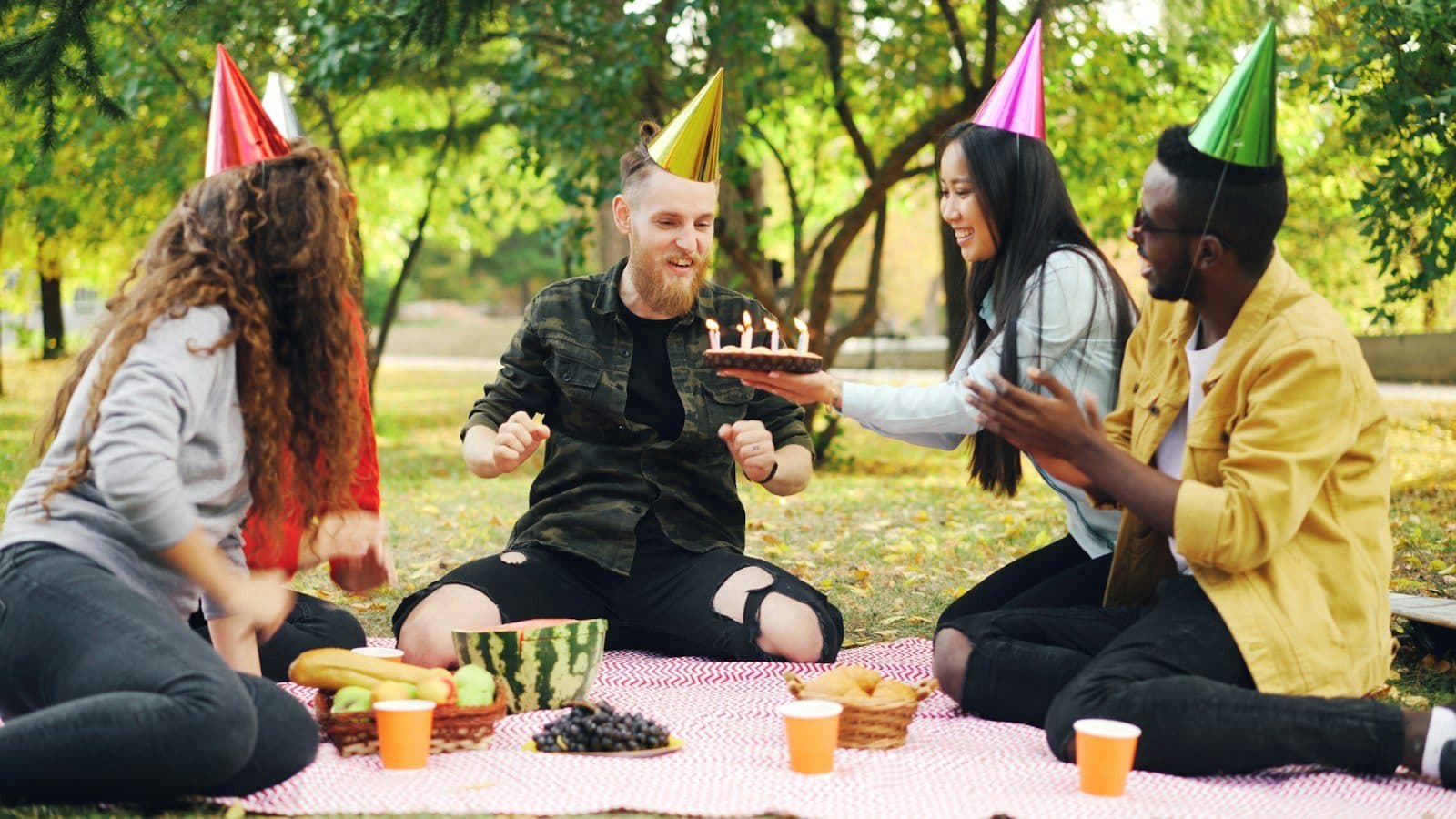 Friends in party hats gathered around a birthday cake on a picnic blanket.